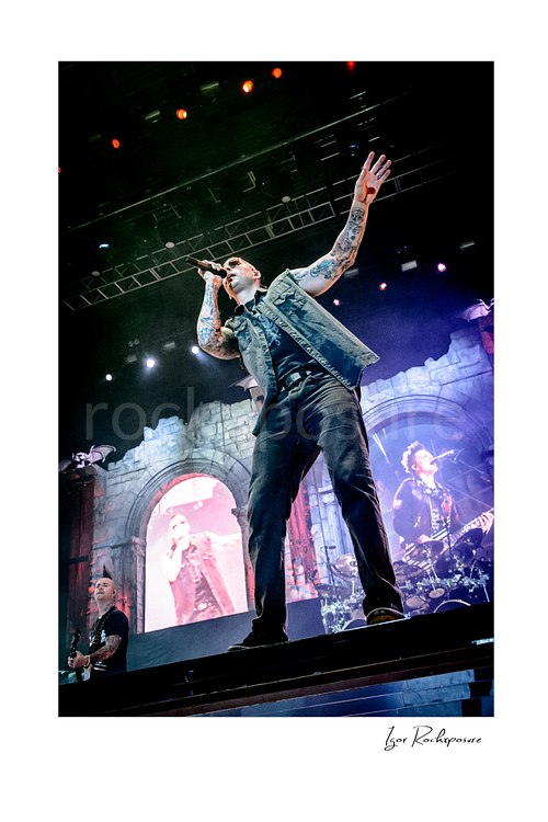Vertical color image of M. Shadows of Avenged Sevenfold singing onstage with one arm raised before glowing screens and dramatic concert lighting