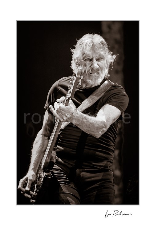 Monochrome concert portrait of Roger Waters playing bass with its neck angled upward before his face, bright light defining his hair and forearms.