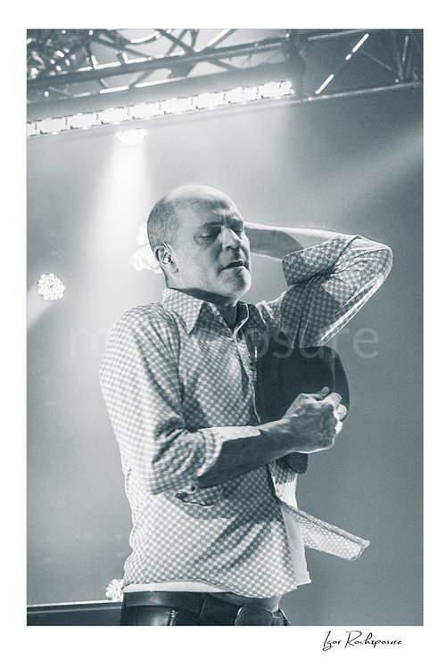 Vertical black and white image of Gord Downie of The Tragically Hip in an emotional live performance moment, holding his hat to his chest with one hand behind his neck under soft stage light