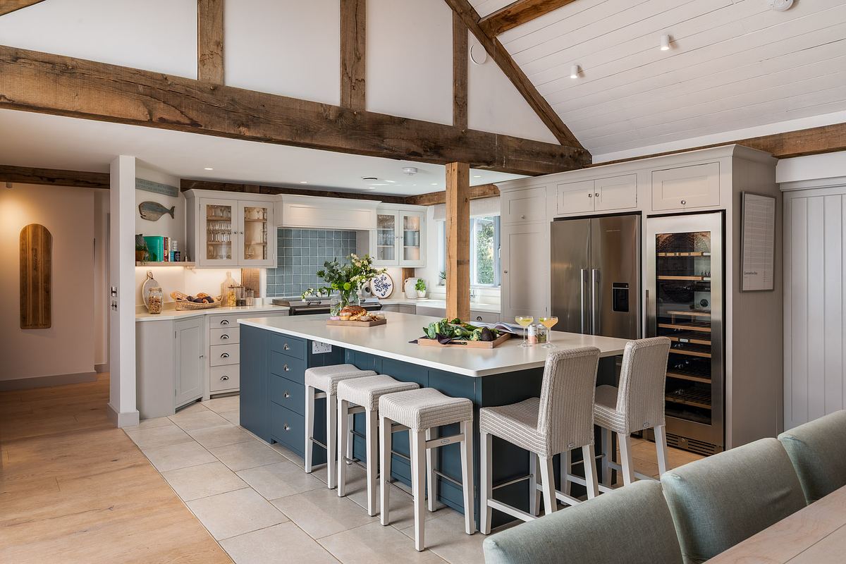 Modern kitchen in a luxury holiday home in Cornwall with exposed wooden beams, large island with bar stools, stainless steel appliances, and open shelving. Light cabinetry and tiled floor create a bright, spacious atmosphere.