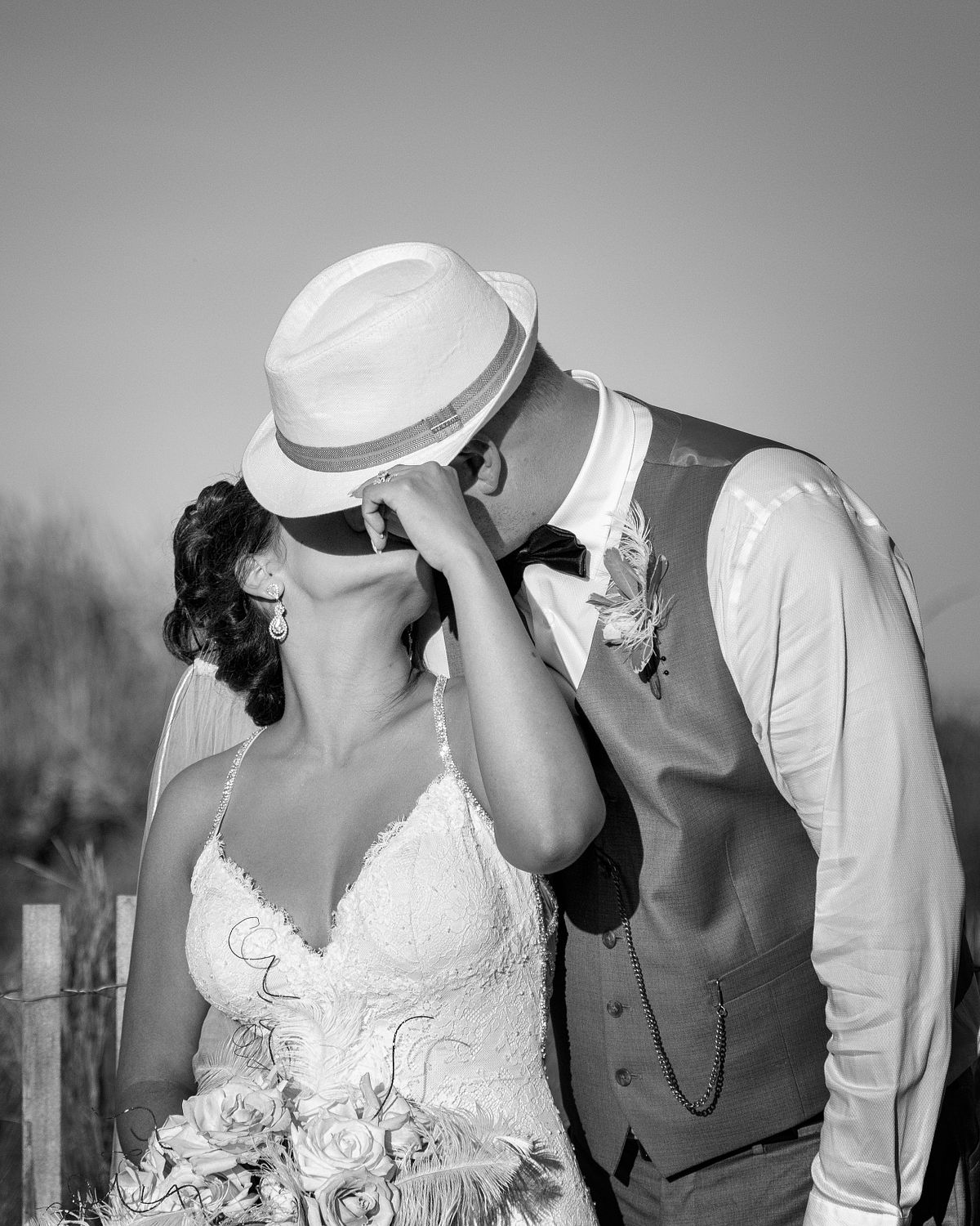 black and white photo of a bride and groom kissing the bride is pulling the groom's hat over her face to make the kiss more private