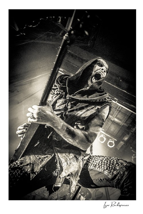 Vertical black and white image of Nergal of Behemoth performing with guitar in a dramatic low-angle close-up with hooded stagewear and corpse paint
