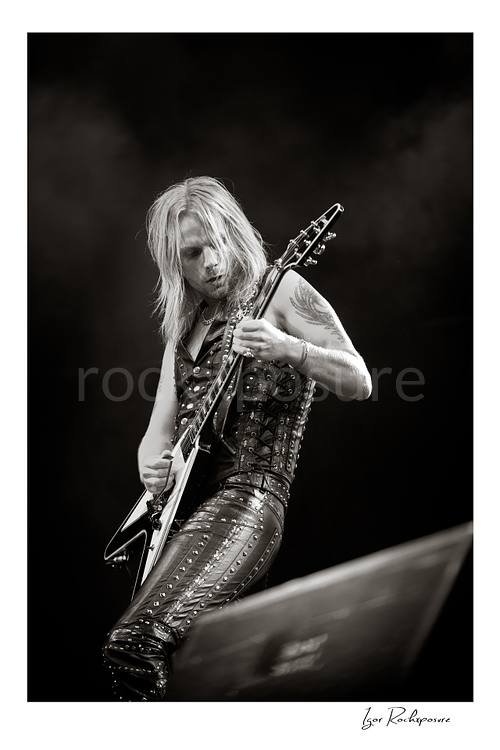 Vertical black and white image of Richie Faulkner of Judas Priest performing live on stage, playing flying V guitar against a dark background