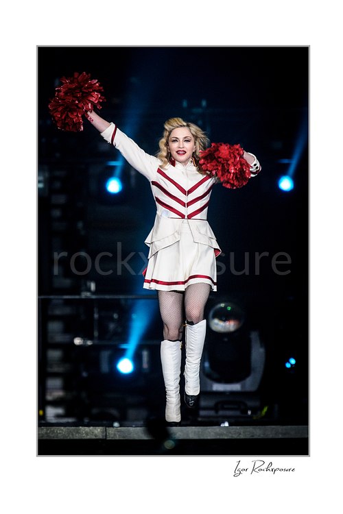 Vertical color image of Madonna performing in a white and red stage outfit while holding red pom-poms under cool blue concert lights