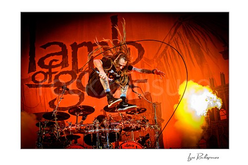 Horizontal color image of Randy Blythe of Lamb of God leaping above the drum kit with flying dreadlocks, microphone cable looping through the air, and a burst of stage fire