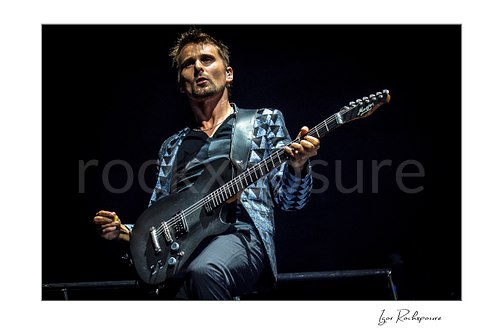 Horizontal color image of Matt Bellamy of Muse playing guitar in a patterned stage jacket against a dark concert background