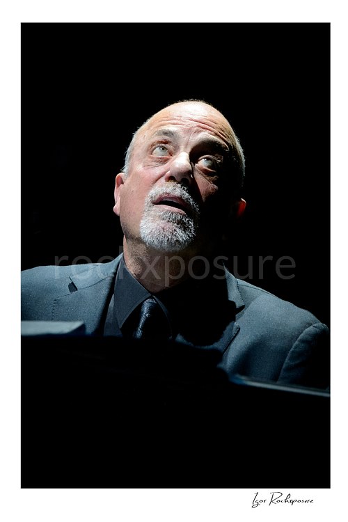 Vertical color image of Billy Joel seated at the piano in a dramatic close-up under a spotlight