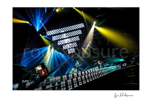 Horizontal color image of Muse performing live on a massive arena stage beneath towering LED screens and vivid blue and yellow spotlights