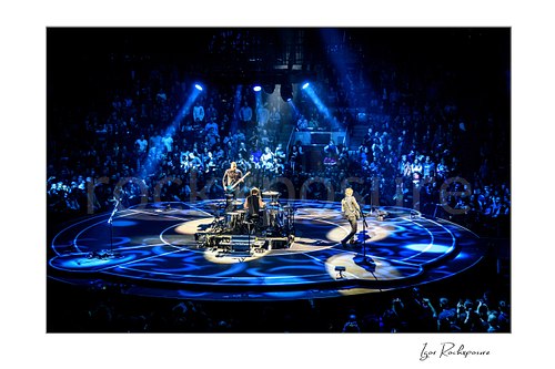 Horizontal color image of Muse performing live on a circular stage under vivid blue spotlights before a packed arena crowd