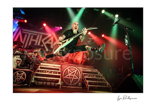 Horizontal color image of Scott Ian of Anthrax performing live on stage, jumping with his guitar in front of the band’s stage set under bright concert lights