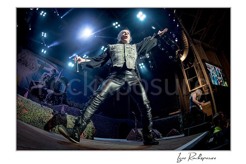Horizontal color image of Bruce Dickinson of Iron Maiden performing live on stage in a dramatic wide stance, holding a microphone under bright concert lights, with a band member visible in the background.