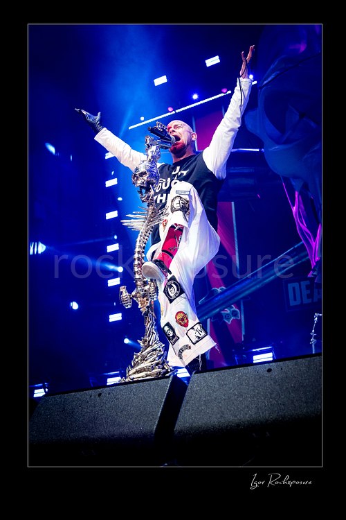 Vertical color image of Ivan Moody of Five Finger Death Punch performing onstage with raised arms beside a skull-covered microphone stand under blue concert light