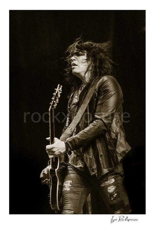 Vertical black and white image of Tom Keifer performing live on stage with a black guitar, hair blowing back in a dramatic concert portrait against a dark background