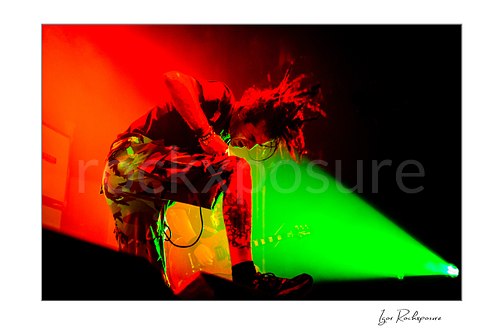 Horizontal color image of Randy Blythe of Lamb of God crouched low on stage with flying hair, handheld microphone, and vivid red and green light