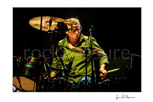 Horizontal color image of Patrick Carney of The Black Keys playing drums under warm stage lighting in a dark concert scene