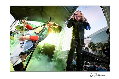 Horizontal color image of D-A-D members Stig Pedersen and Jesper Binzer performing live on stage with stage smoke and bright backlighting