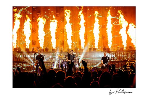 Horizontal color image of Nickelback performing onstage before a packed crowd with towering fire columns and bright concert beams
