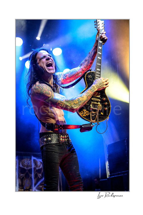 Vertical color image of Chuck Garric of Beasto Blanco performing live shirtless with a black guitar, mouth open in a fierce expression under vivid blue and yellow stage lights