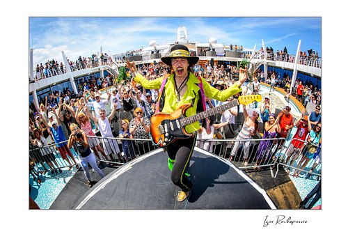 Horizontal color image of Randy Hansen playing guitar at the front of an outdoor ship deck stage with a large cheering crowd behind him