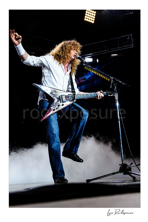 Vertical color image of Dave Mustaine of Megadeth singing and playing guitar on stage with one leg raised above low stage smoke