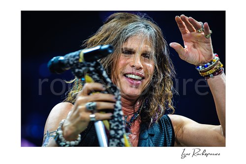 Horizontal color image of Steven Tyler of Aerosmith performing live on stage with a scarf-wrapped microphone stand, raised hand, bracelets, and tousled hair
