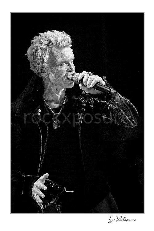 Vertical black and white image of Billy Idol singing into a microphone in a tight live performance portrait