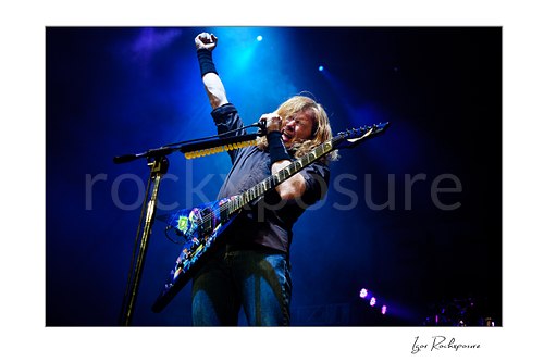 Horizontal color image of Dave Mustaine of Megadeth singing into a microphone with one arm raised and guitar under blue stage lights
