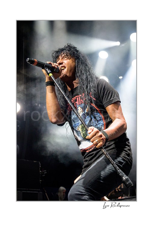 Vertical color image of Joey Belladonna of Anthrax performing live on stage, singing into a microphone under bright concert lights