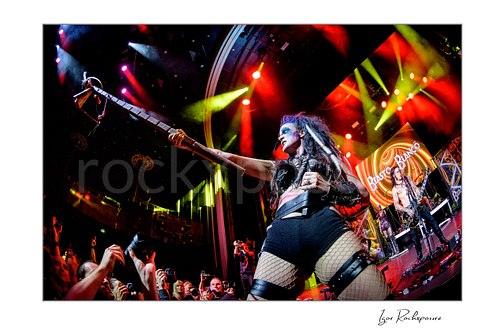 Horizontal color image of Calico Cooper of Beasto Blanco performing live at the front of the stage, extending a long stage prop toward the crowd under vivid red and yellow lights with Chuck Garric visible behind her