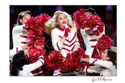 Horizontal color image of Madonna singing into a microphone in a white and red stage outfit surrounded by dancers with bright red pom-poms