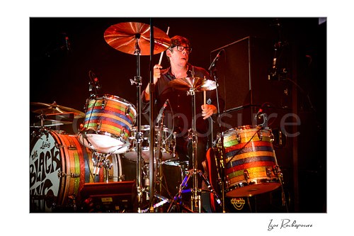 Horizontal color image of Patrick Carney of The Black Keys playing drums behind a striped drum kit under warm red stage lighting