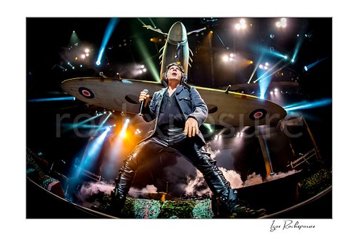 Bruce Dickinson of Iron Maiden performing live on stage beneath a large airplane stage prop, holding a microphone and standing in a wide pose under bright concert lights.