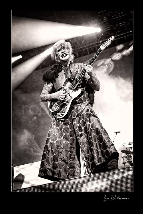 Vertical black and white image of John 5 performing live on stage with an electric guitar, theatrical makeup, patterned stage outfit, and bright backlighting