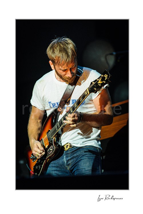 Vertical color image of Dan Auerbach of The Black Keys playing guitar under warm stage lighting in a dark concert scene