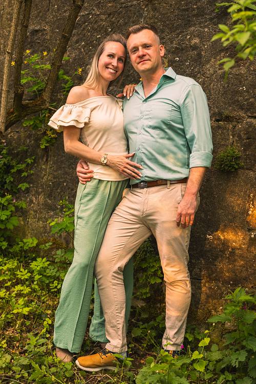 Ein lächelndes Paar steht eng umschlungen vor einer rustikalen, bewachsenen Steinwand. Die Frau trägt ein helles Off-Shoulder-Top und eine grüne Hose.