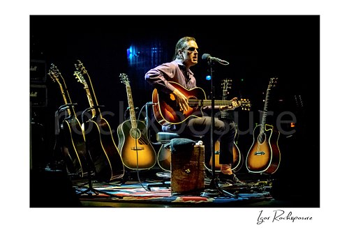 Horizontal color image of Joe Bonamassa seated on stage with an acoustic guitar, singing into a microphone and surrounded by multiple guitars