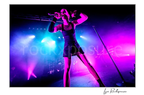 Horizontal color image of Shirley Manson singing into a microphone during a Garbage concert in a dramatic full-stage view under vivid pink and blue lighting