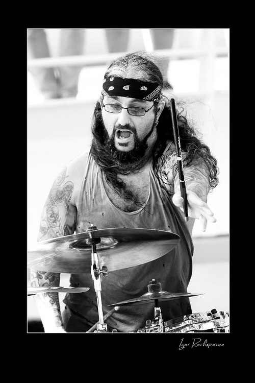 Vertical black and white image of Mike Portnoy at the drum kit with a drumstick extended forward and an intense expression