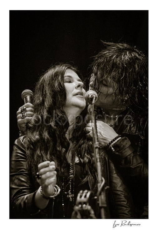Vertical black and white image of Tom Keifer and Savannah sharing the microphone in a close live performance moment, framed tightly together at center stage