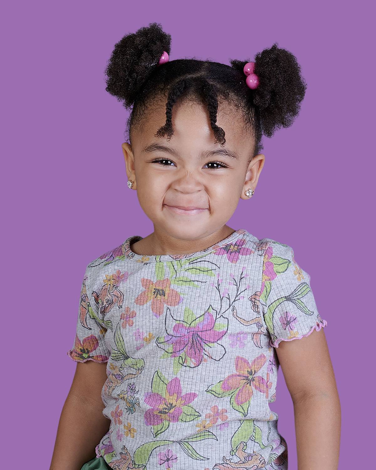 A professional school portrait by ProClicks Photography of a smiling young girl with curly hair styled in two puffs with pink beads, wearing a grey floral t-shirt against a solid purple background.
