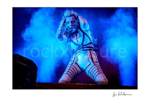 Horizontal color image of Maria Brink of In This Moment kneeling on stage in a dramatic live performance pose, framed by dense blue stage smoke and intense blue lighting