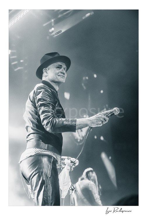 Vertical black and white image of Gord Downie of The Tragically Hip holding a microphone on stage, wearing a hat and leather jacket with bright concert haze and a blurred band member behind him