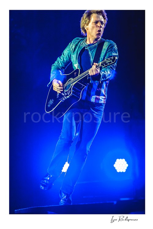 Vertical color image of Jon Bon Jovi performing live with a black acoustic guitar under vivid blue stage lighting