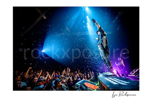 Horizontal color image of Alice Cooper performing live on stage on a runway above the crowd with blue spotlights, raised arm, and floating bubbles