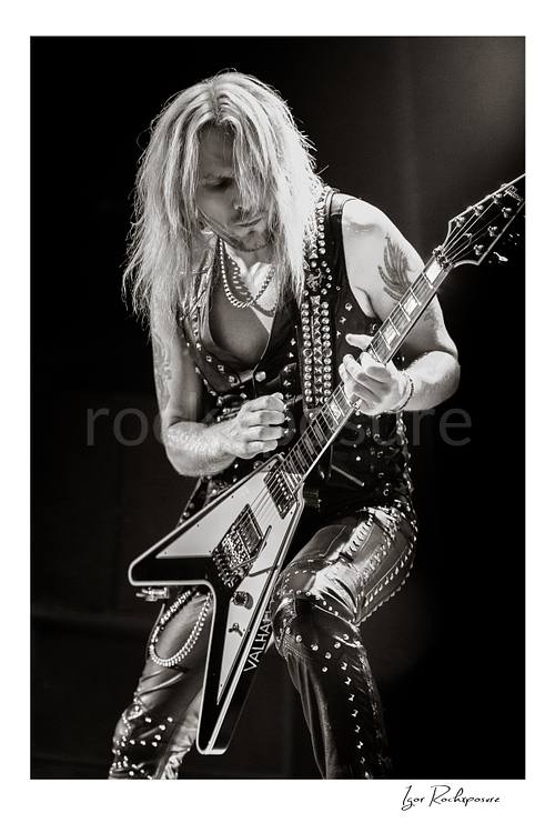 Vertical black and white image of Richie Faulkner of Judas Priest performing live on stage, playing flying V guitar under dramatic concert lighting