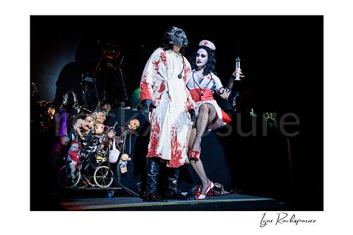 Horizontal color image of Alice Cooper performing live on stage in a blood-stained straitjacket beside his wife dressed as a nurse holding a large syringe