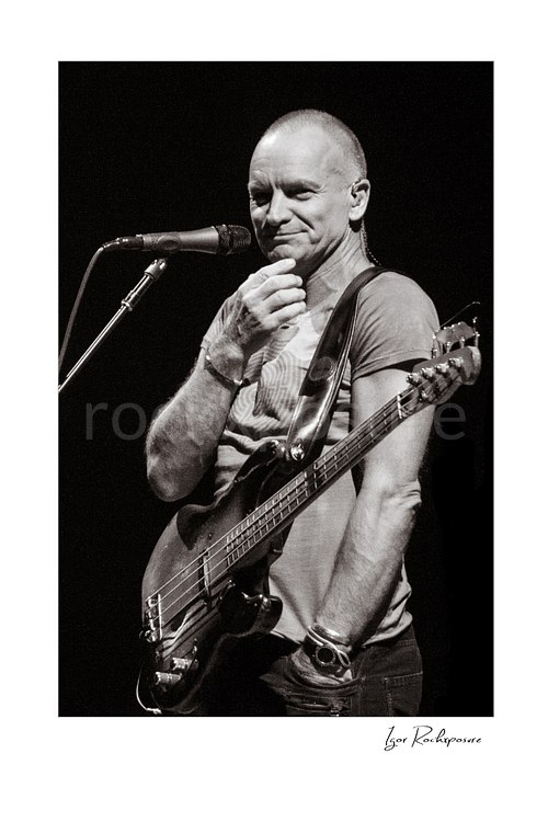 Vertical black and white image of Sting holding a bass guitar beside a microphone in a quiet live performance portrait