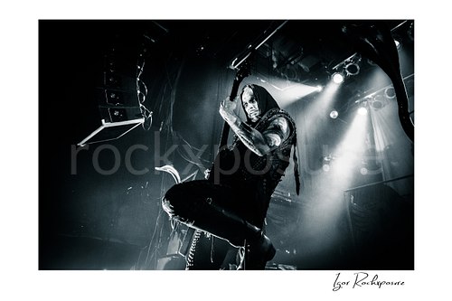 Horizontal black and white image of Nergal of Behemoth performing with guitar in a dramatic low-angle stage shot under bright concert lights