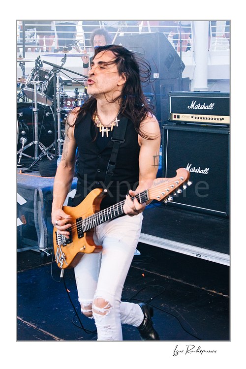 Vertical color image of Nuno Bettencourt performing live on stage with an electric guitar, windblown hair, and ripped white jeans