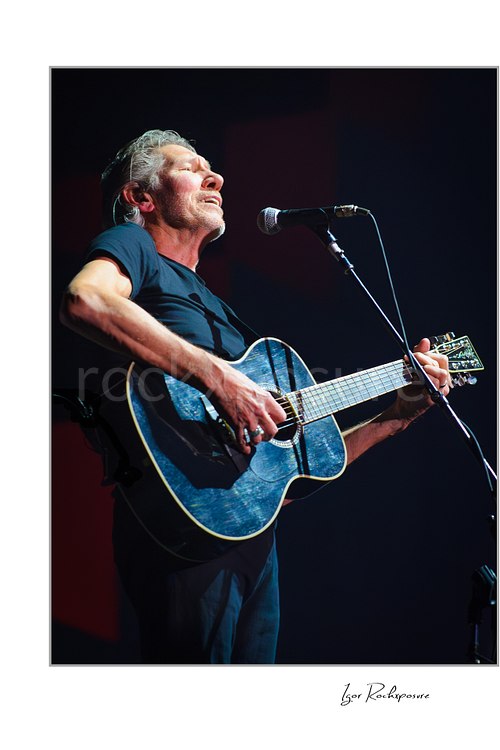 Roger Waters sings with his eyes closed at a stand microphone while playing a dark acoustic guitar reflecting blue light against a dim background.